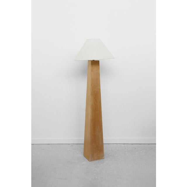 Floor lamp defined by a tall, monolithic column base in a blonde wood finish, paired with a simple tapered shade. The form...
