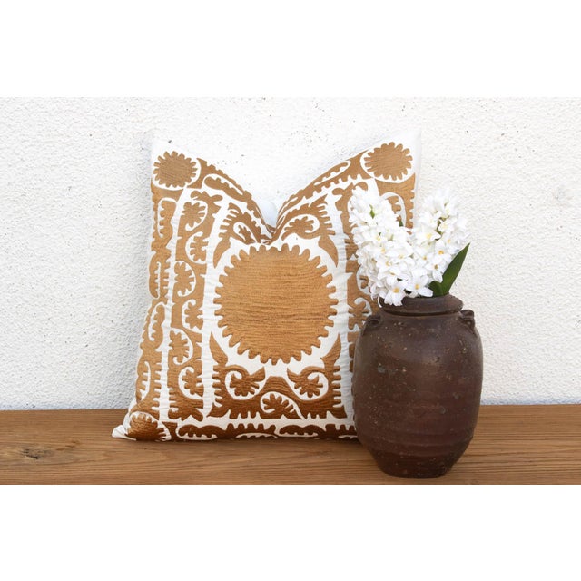 A dense arrangement of gold-tan motifs covers nearly the full face of this pillow — a sun-disc medallion at center, framed...
