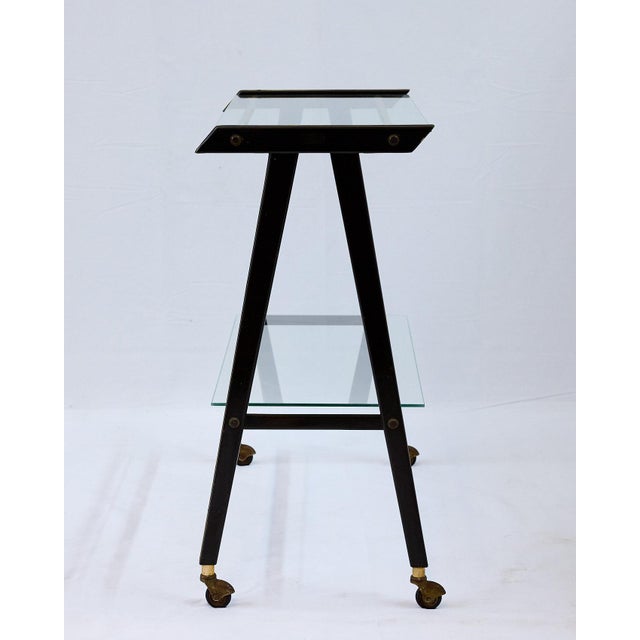 Mid-century Italian bar trolley or vintage tv cart having a black, brass-inlaid frame with brass cross stretchers that...