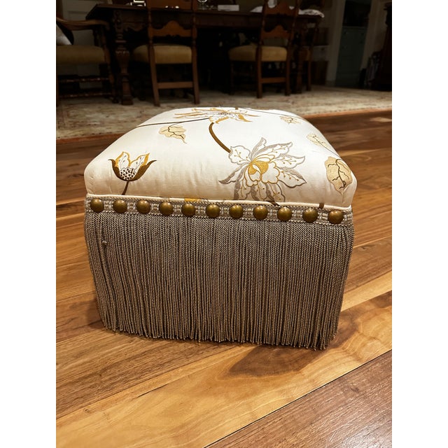 Tassle-fringed upholstered footstool with studded trim. Cream silk floral embroidery pattern.