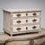 Cream-painted miniature commode with three drawers, rounded fluted corners, and small baluster feet. The bronze fittings...