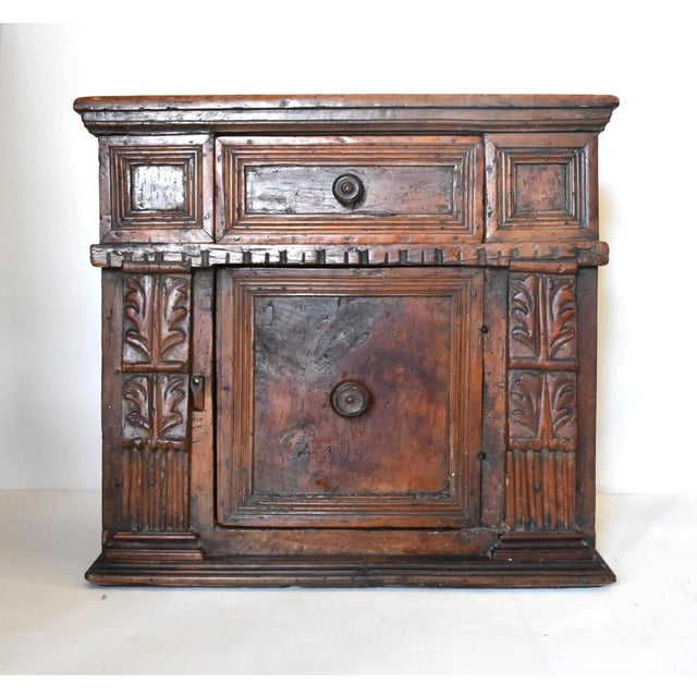 An antique Italian credenzino or small cabinet with one door and one drawer, hand-hewn and carved of heavy walnut with...