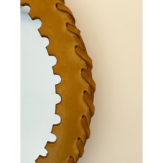 A round mirror encircled by a wide band of tan leather, entirely handcrafted, adorned with lacing and a scalloped border....