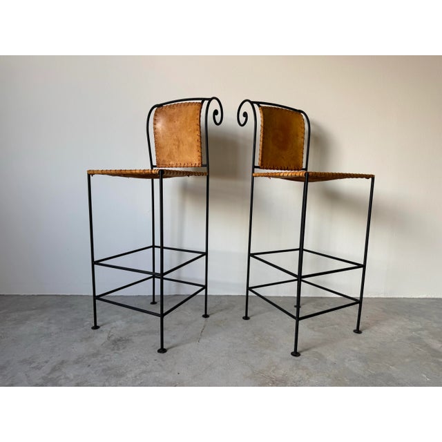 Beautiful pair of vintage Spanish-style barstools featuring hand-forged wrought iron frames and hand-stitched natural...