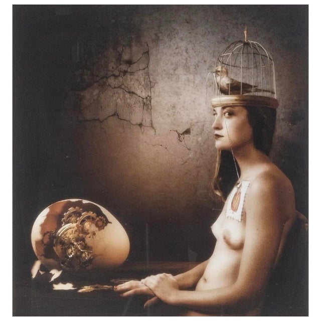 A woman sits with a birdcage crowning her head — a live bird perched inside it — while beside her an ostrich egg has...