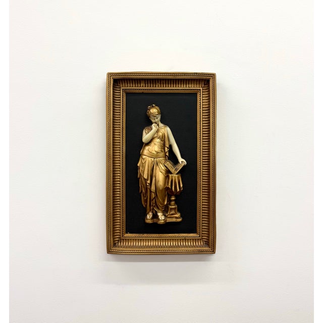 A decorative wall plaque featuring a raised classical female figure finished in warm gilt tones against a black textured...