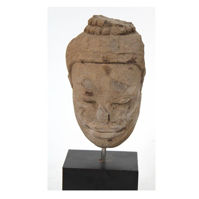 A small carved and weathered sandstone Buddha head in tan-brown tones with darker mineral staining, downcast, meditative...