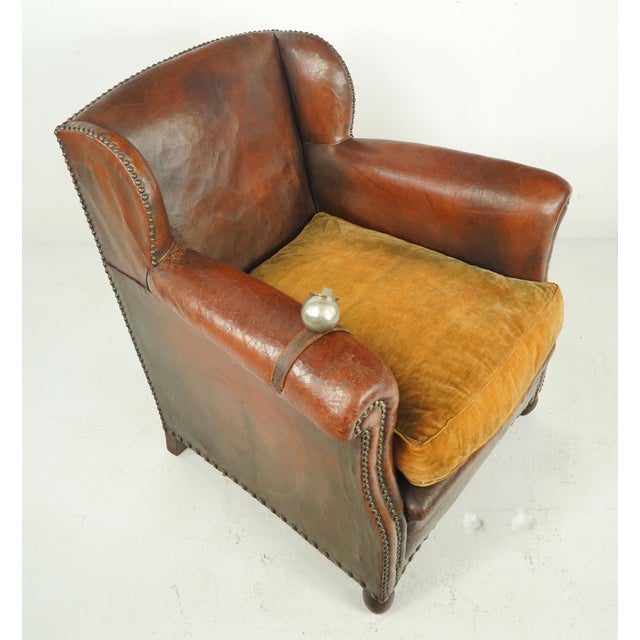 Vintage dark brown leather studded club chair made in France. It has a plush amber cushioned seat, studded details, and...