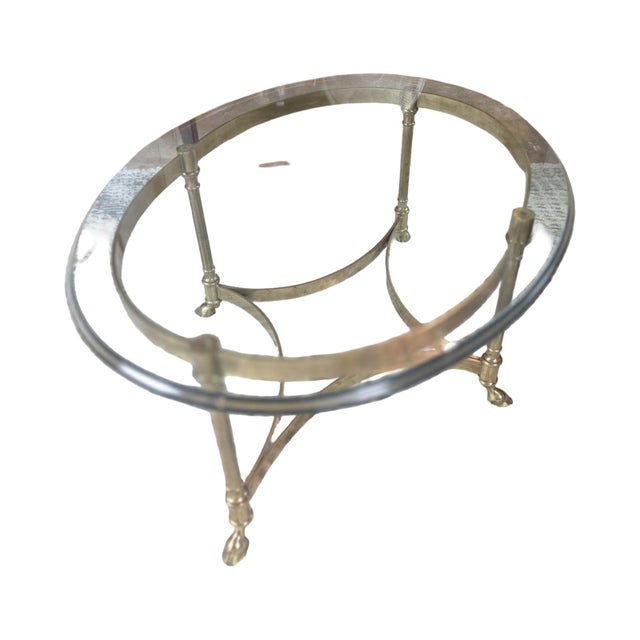 Elegant vintage oval coffee table featuring a substantial beveled glass top supported by a sculptural brass base. The...
