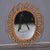 An oval mirror framed in bent rattan, looped into a scalloped, sunburst-style pattern that fans out around a woven rattan...