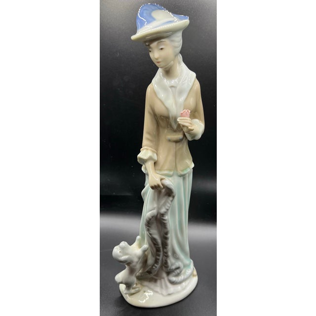 This item is a vintage porcelain figurine of a woman holding a flower with a puppy at her feet, identified in the style of...