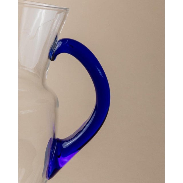 This vintage glass jug, crafted in the 1970s in Italy, features clear glass with a striking blue handle, making it a...