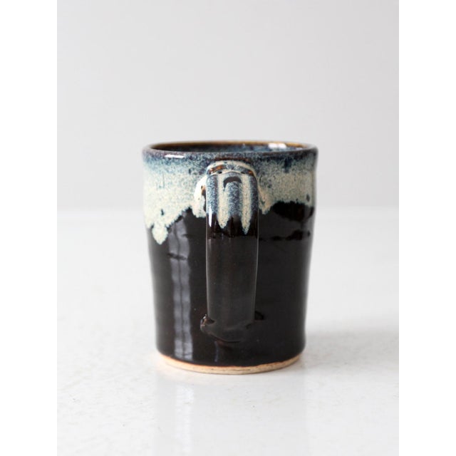 A vintage handmade studio pottery mug in black with a soft blue drip glaze along the rim. The gentle variation in tone...