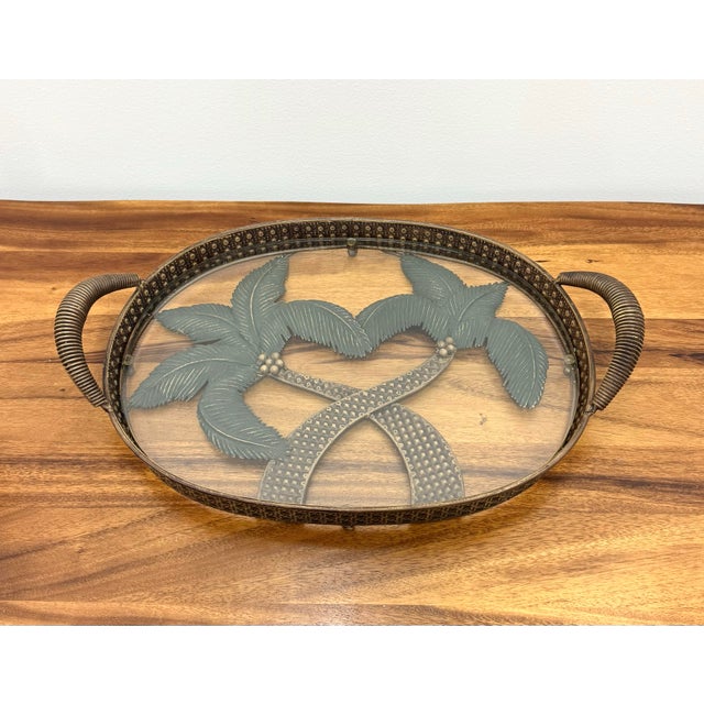 A charming vintage serving tray featuring an oval glass surface showcasing a pair of stylized crossed palm trees beneath...