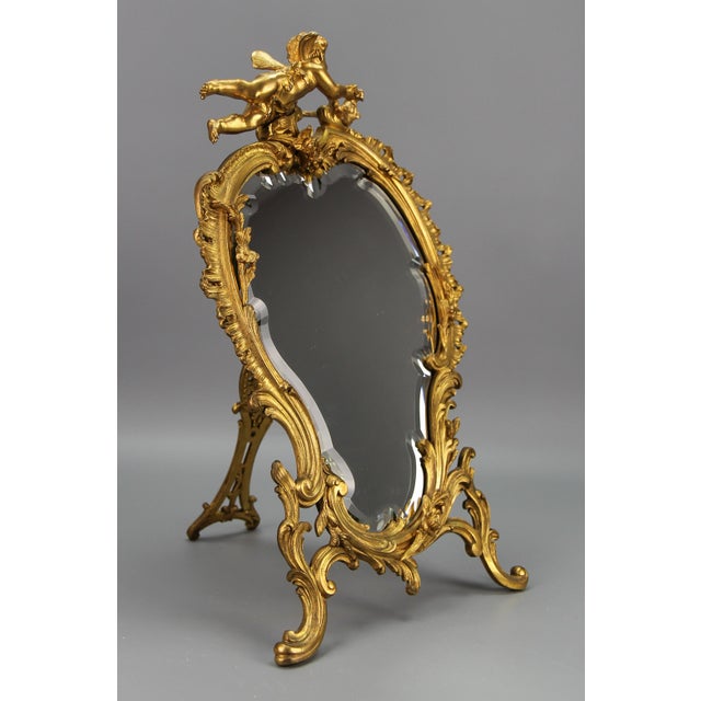 Antique French gilt bronze Rococo style desktop mirror with cherub and bird from the early 20th century. Adorned with...