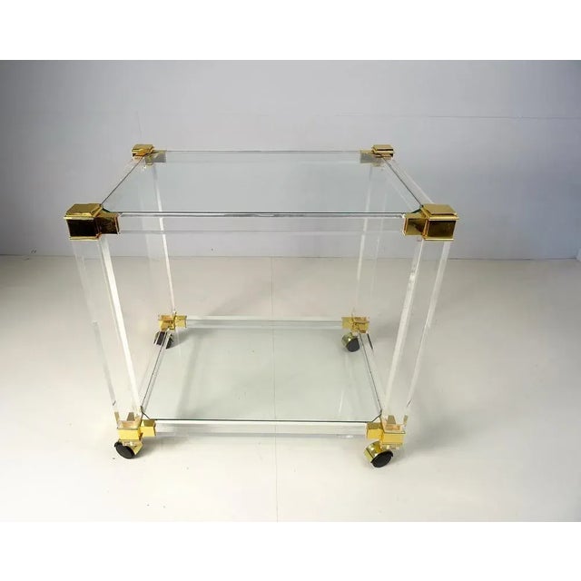 This bar cart is made of acrylic glass with two tiers and golden details. The bar cart features four wheels and two glass...