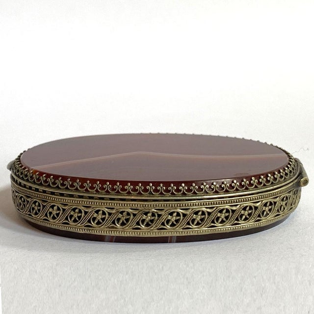 DETAILS An antique, oval box with a polished, banded agate stone, hinged lid and base. With mirrored interior and brass...