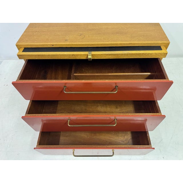 Mid-century chest of drawers by Heal’s, featuring a striking contrast of warm wood grain and red lacquered drawer fronts....