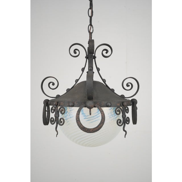 Spanish Revival style pendant light featuring a heavy wrought iron frame with ornate scrollwork. The centerpiece is a hand...