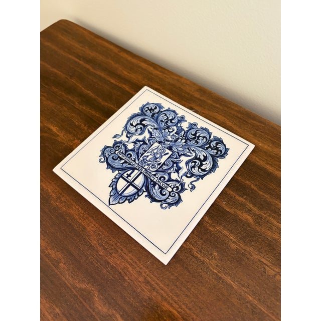 A classic Dutch Delft-style hand painted blue and white porcelain tile featuring an intricately rendered heraldic coat of...