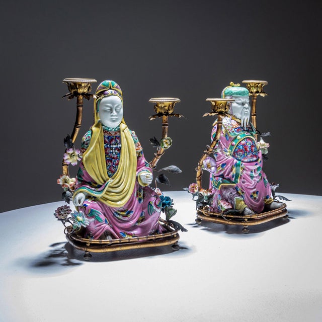 A pair of Famille Rose table candlesticks with a male and female chinoiserie figure sitting on a plinth in the shape of...