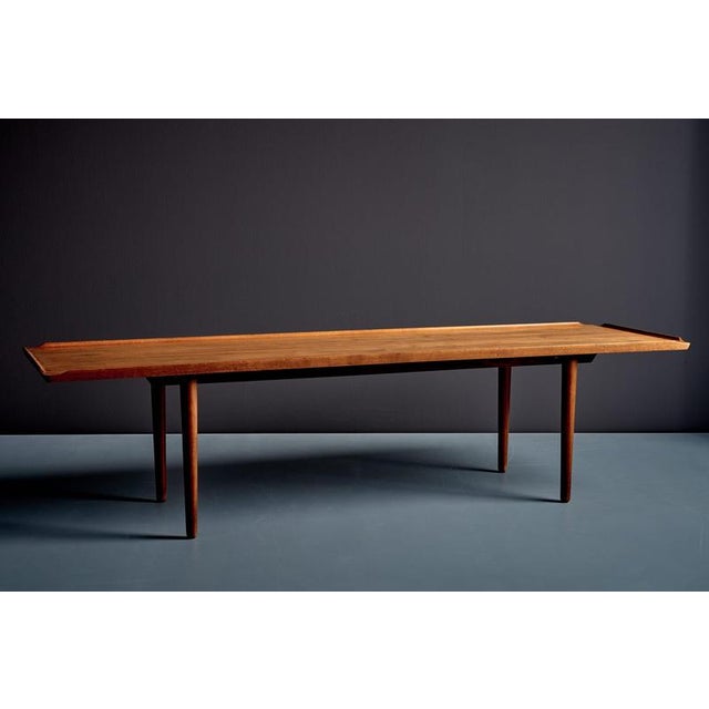 Danish coffee table from the 1960s, featuring a long rectangular teak top with a gently raised, shaped edge. The slender,...