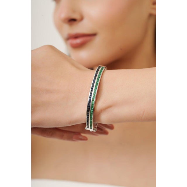 Elegant bangle crafted in 925 sterling silver, featuring a sleek channel-set design with vibrant green emeralds and deep...