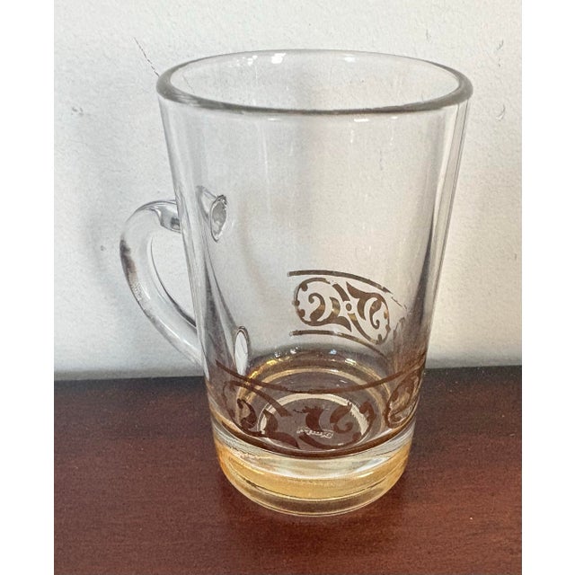 Elegant set of seven vintage Italian glass mugs with gold enamel scroll decoration and weighted amber-tinted bases. Each...