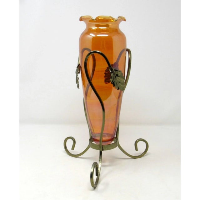 A vintage amber hand-blown carnival glass amphora-style vase set in an ornate metal tripod stand. The vase has a ruffled...