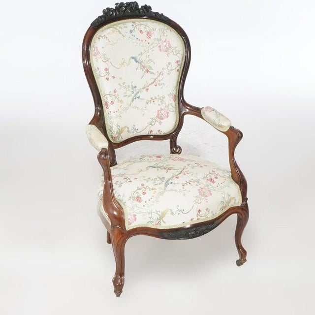 An antique pair of Victorian parlor armchairs offer rosewood construction with carved floral crest over upholstered seat,...