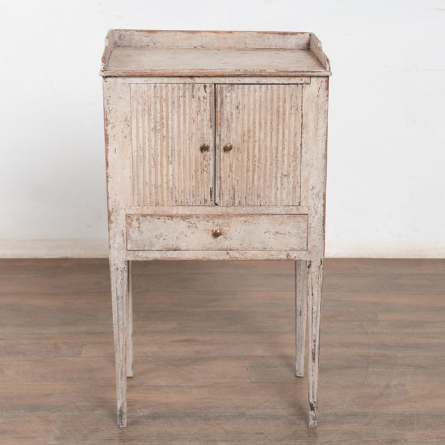 Swedish country-style nightstand or small cabinet in pine with fluted doors and tapered, fluted legs. The aged grey paint...