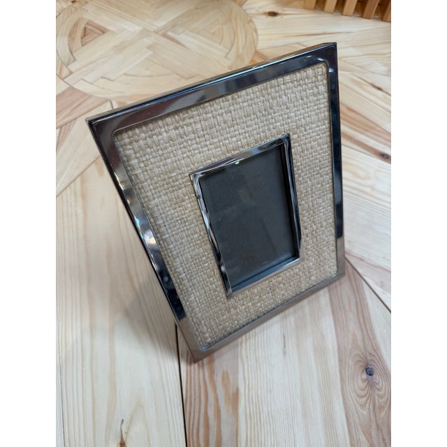 A tabletop picture frame with a natural woven rattan surround and polished silver-tone metal borders. The contrast of...