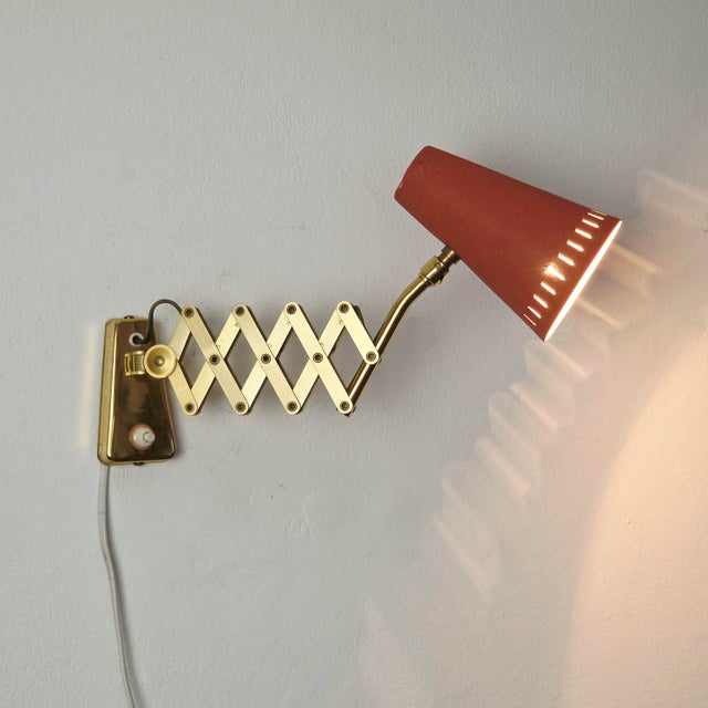An elegant, adjustable vintage scissor wall lamp from the 1950s features a brass frame and a coral-red lacquered aluminum...