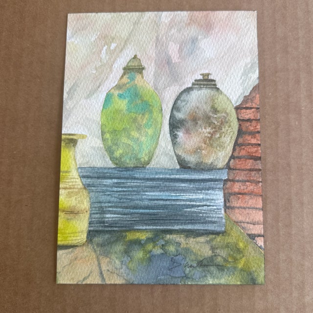 Rich with texture and earthy tones, this watercolor still life showcases aged pottery resting on a rustic surface. The...