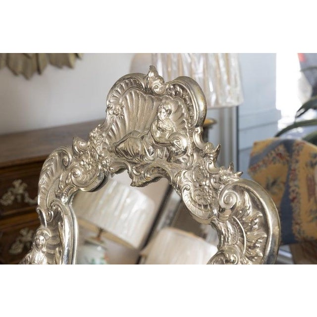 An exceptional Continental repoussé silvered table mirror featuring an exuberant Rococo-inspired frame richly decorated...