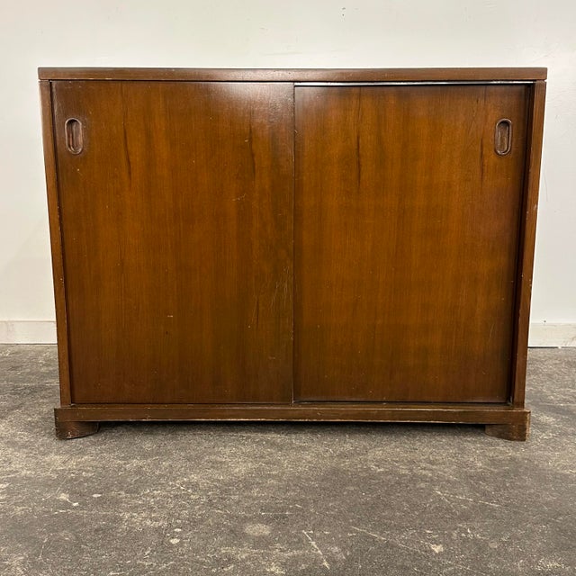 Mid Century John Widdicomb walnut sideboard or storage chest with sliding doors. The sliding doors feature recessed oval-...