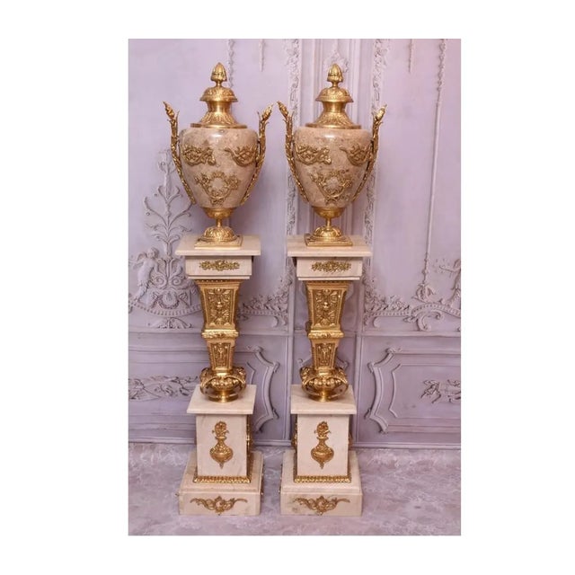A magnificent pair of monumental Neoclassical-style beige marble urn amphorae set atop matching marble pedestal columns....