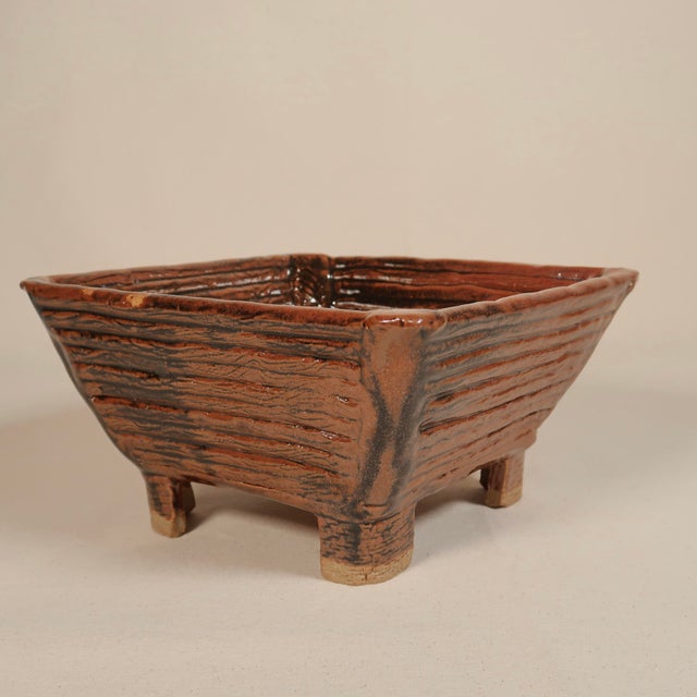 This handcrafted ceramic vessel features a square, tapered form elevated on four short, block-like feet. The vessel is...
