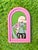 This artwork depicts a stylized figure with a pale, greenish complexion and a playful yet haunting expression, holding a...