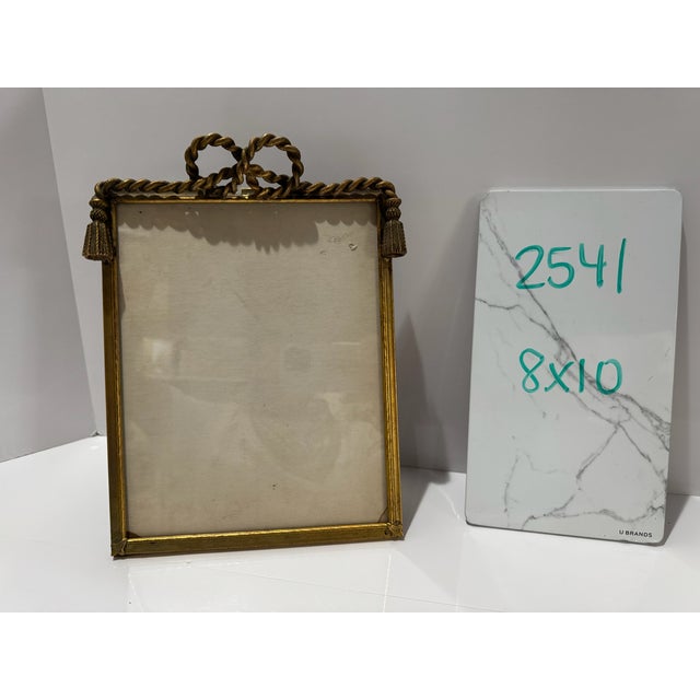 Decorative gilt metal frame with twisted rope border, ribbon bow crest, and tassel details. Measures approximately 8 x 10...