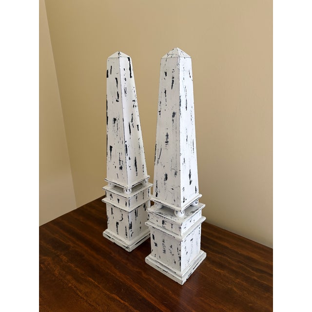 A pair of painted wood obelisks, rendered in a deliberately timeworn finish that evokes architectural fragments softened...