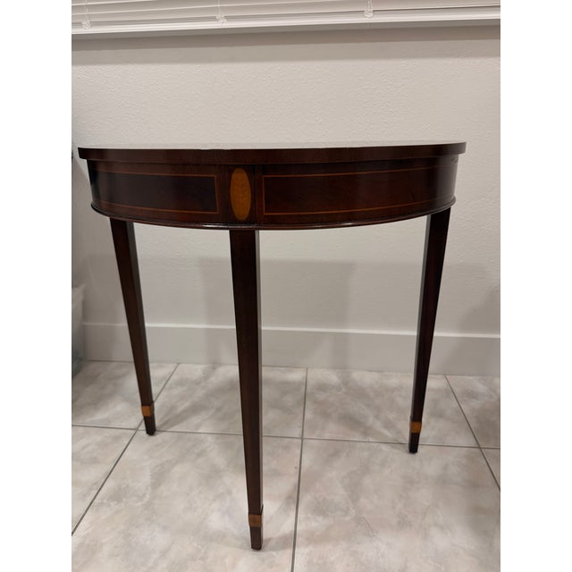 An elegant Federal style demilune console table by the renowned Baker Furniture Company. Authentic Baker Furniture piece...