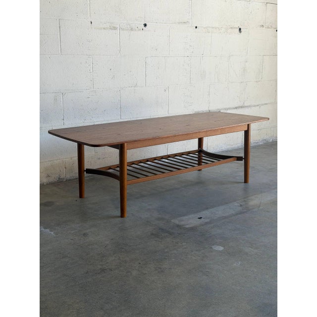 This rectangular teak coffee table combines a long narrow top with a full-length slatted lower shelf. Rounded corners and...