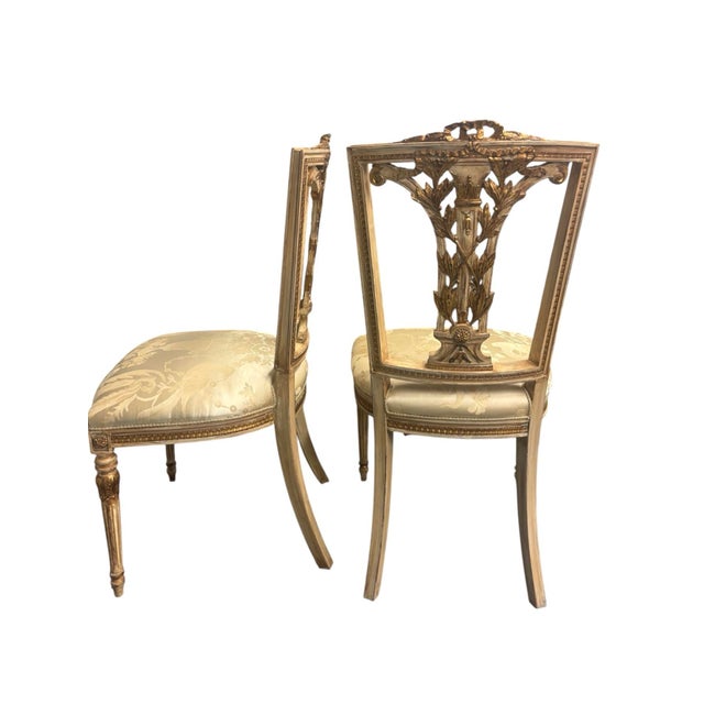 Cream wood chairs with gold detailing carved into the back. Seat is covered in a shiny ivory fabric with a subtle floral...