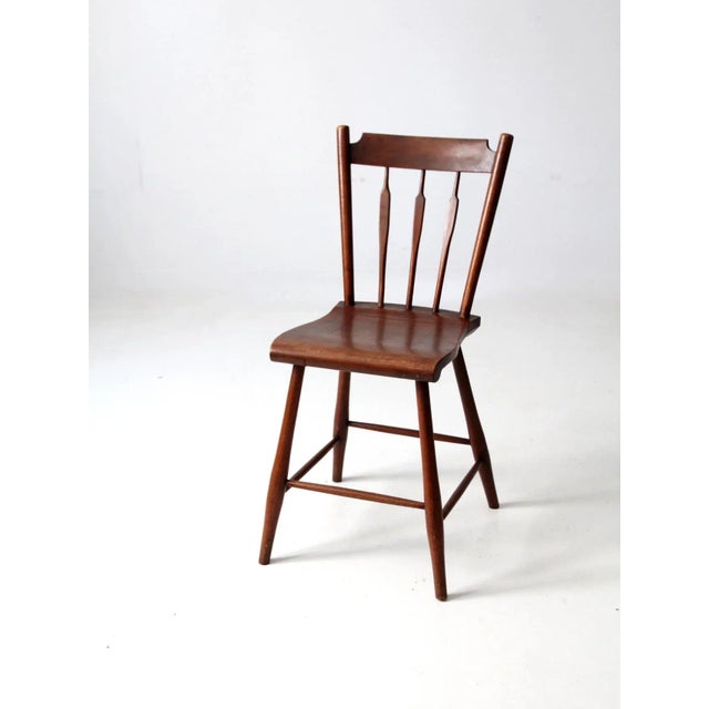 An antique Windsor-style accent chair featuring a decorative spindle back, a shaped wooden seat, and gracefully splayed...