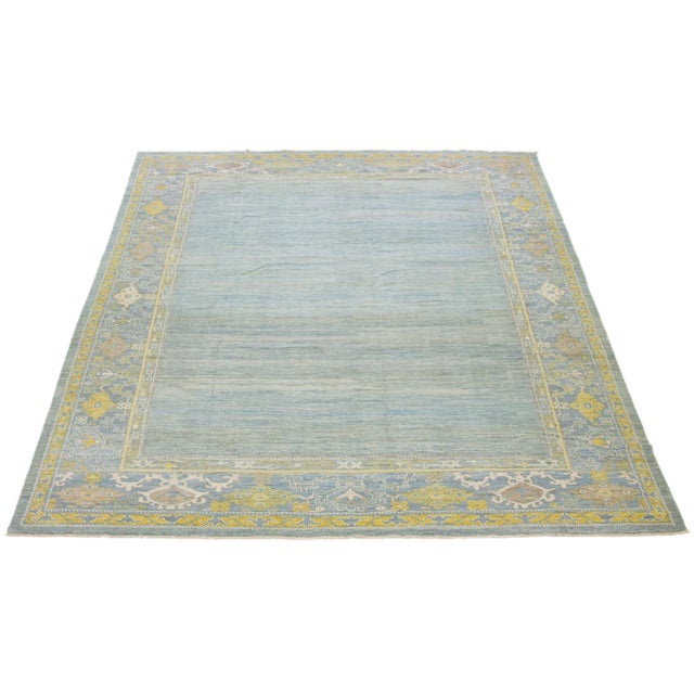 This hand-knotted wool rug features a light blue background with yellow and beige accents in a framed design. This rug...