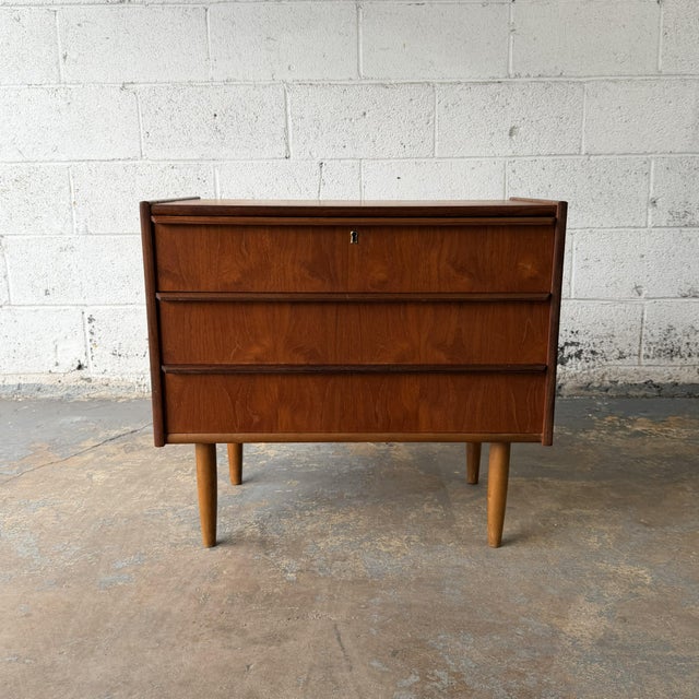 A Danish teak three-drawer chest from the 1960s with slim integrated pulls, tapered legs, and a compact raised case. The...