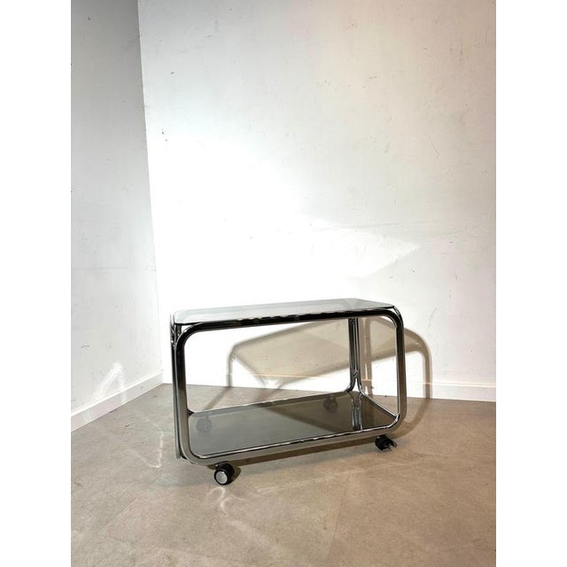 Elegant vintage serving trolley or side table featuring a sleek chrome tubular frame combined with smoked glass shelves....