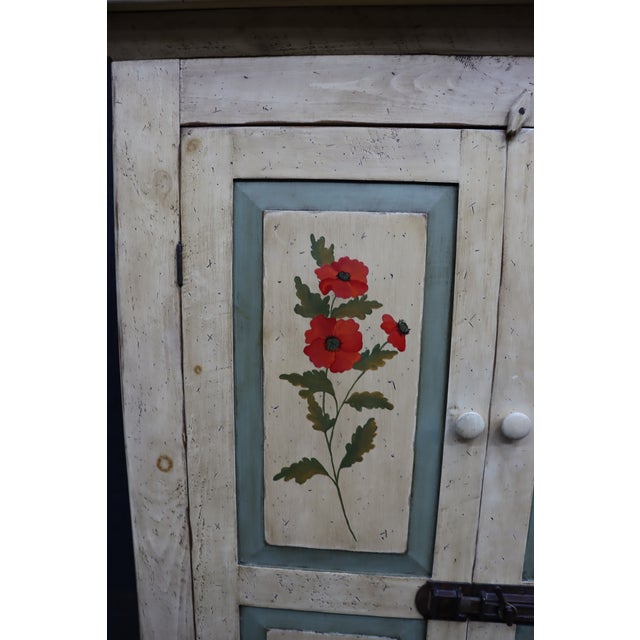 Pine linen press featuring a distressed cream-painted finish with muted blue-green accents and hand-painted floral panels...