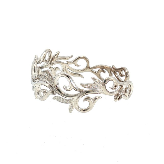An 18k white gold cuff bracelet with intricate swirling vine motifs. Adorned with 80 brilliant-cut white diamonds totaling...